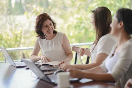 Smiling woman in a white top talks with colleagues over laptops and a notebook in a bright, authentic team portrait.