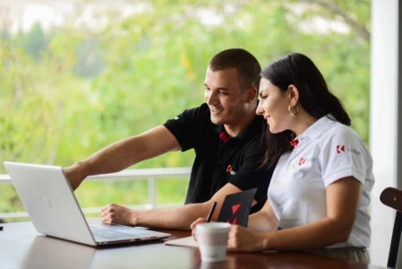 Two smiling colleagues in branded polos examine a laptop together in a natural light team portrait, blurred green background.