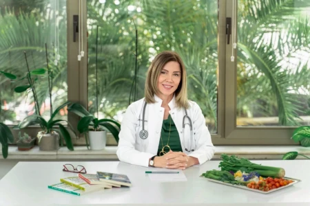 Smiling doctor in a white coat with stethoscope seated at a table by a leafy window, authentic natural professional headshot.