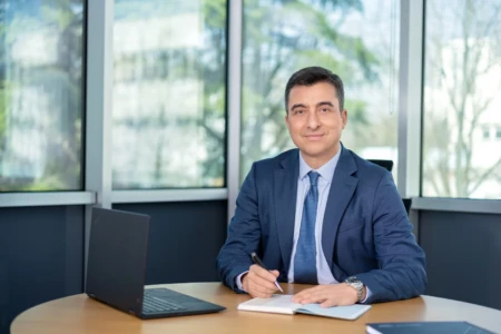 Smiling man in a navy suit sits at a desk writing in a notebook by windows, confident business headshot with natural light.