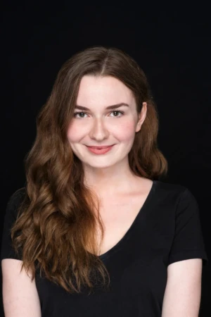 Smiling young woman in black shirt posed against a dark backdrop, studio-lit actor headshot with an authentic, confident look.
