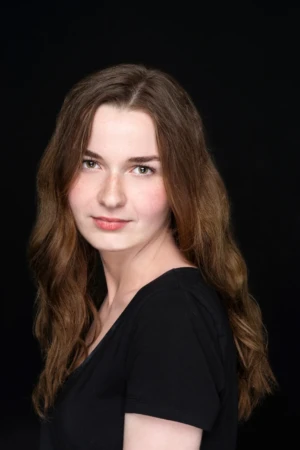 Studio-lit actor headshot of a young woman in a black top with wavy brown hair, soft smile and dark seamless background.
