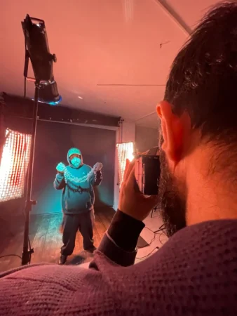 Photographer frames a hooded man holding chains under teal and magenta studio lights in a cinematic studio portrait.