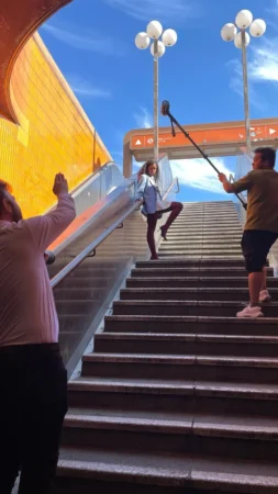 Woman in a blazer poses confidently on outdoor metro stairs against an orange wall and blue sky, outdoor portrait in Berlin.