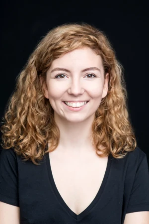 Smiling woman with curly hair against a black background wearing a V-neck — professional headshot in studio light.