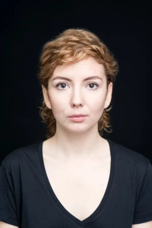 Frontal studio-lit professional headshot of a young woman with short wavy hair, neutral expression, black background.
