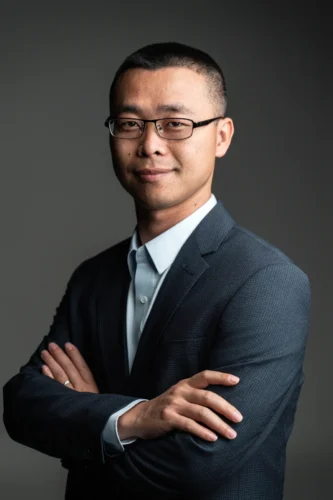 DSC03589 Selbstbewusstes Business-Foto eines Mannes mit Brille und dunklem Anzug, der die Arme vor einem weichen, grauen Hintergrund mit Studiolicht verschränkt.