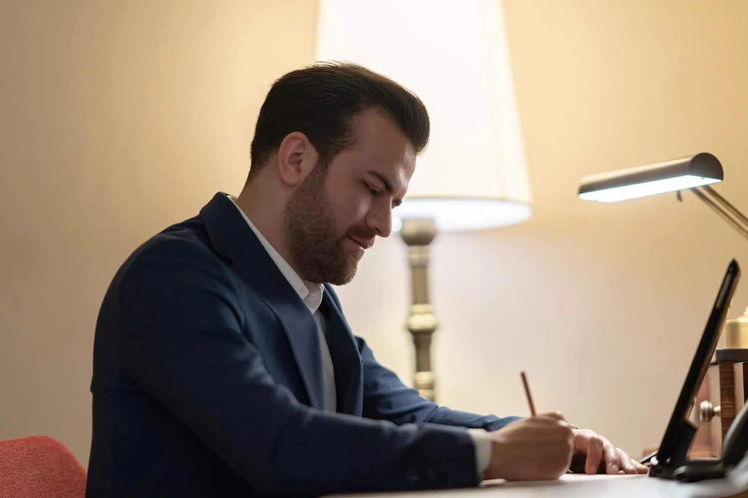 Personal Branding Portrait - Professionelles Schreiben im modernen Arbeitsumfeld Ein Mann in einem marineblauen Blazer schreibt nachdenklich, beleuchtet von einer Schreibtischlampe, mit einem Tablet und einem gemütlichen Ambiente im Hintergrund.
